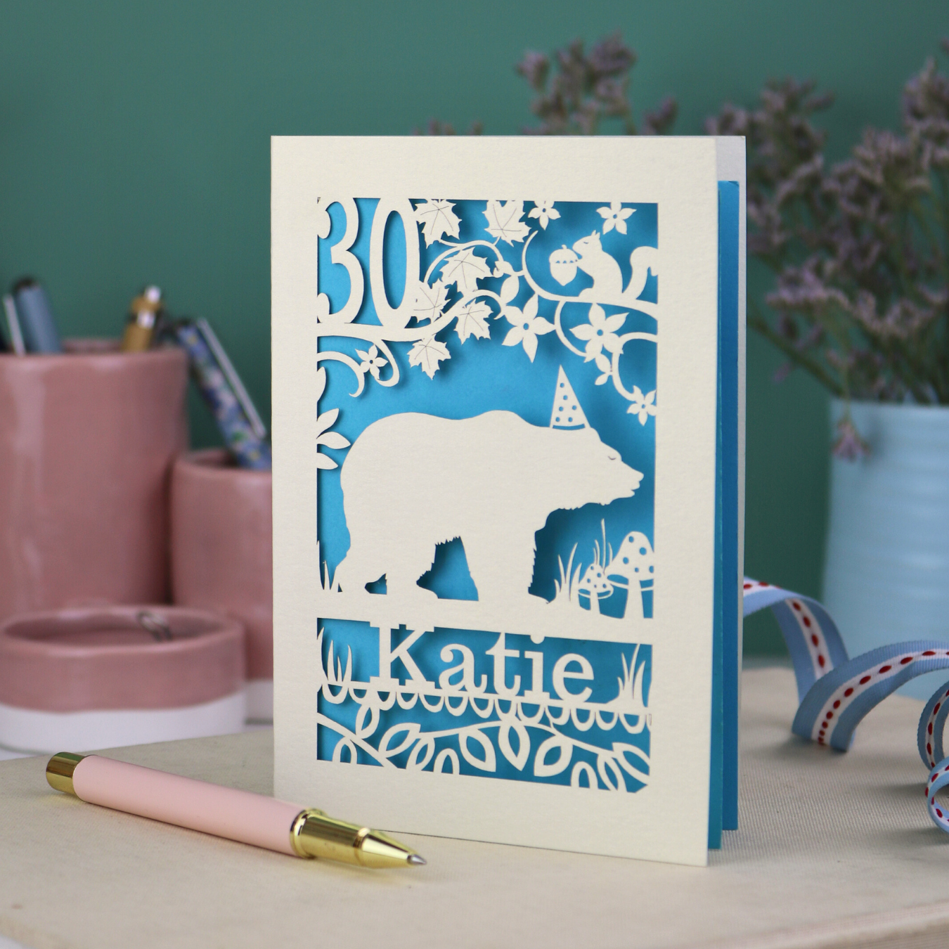 A laser cut birthday card personalised with an age and name, and featuring a cut out bear in a party hat with woodland border.