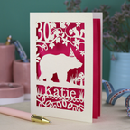 A laser cut birthday card personalised with an age and name, and featuring a cut out bear in a party hat with woodland border.