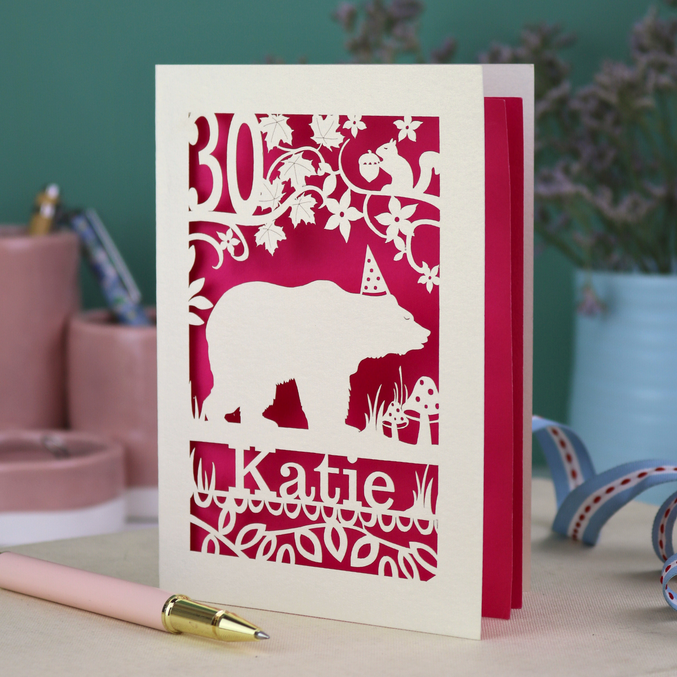 A laser cut birthday card personalised with an age and name, and featuring a cut out bear in a party hat with woodland border.