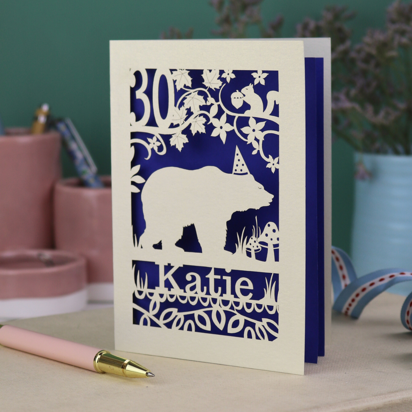 A laser cut birthday card personalised with an age and name, and featuring a cut out bear in a party hat with woodland border.
