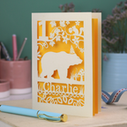 A laser cut birthday card personalised with an age and name, and featuring a cut out bear in a party hat with woodland border.