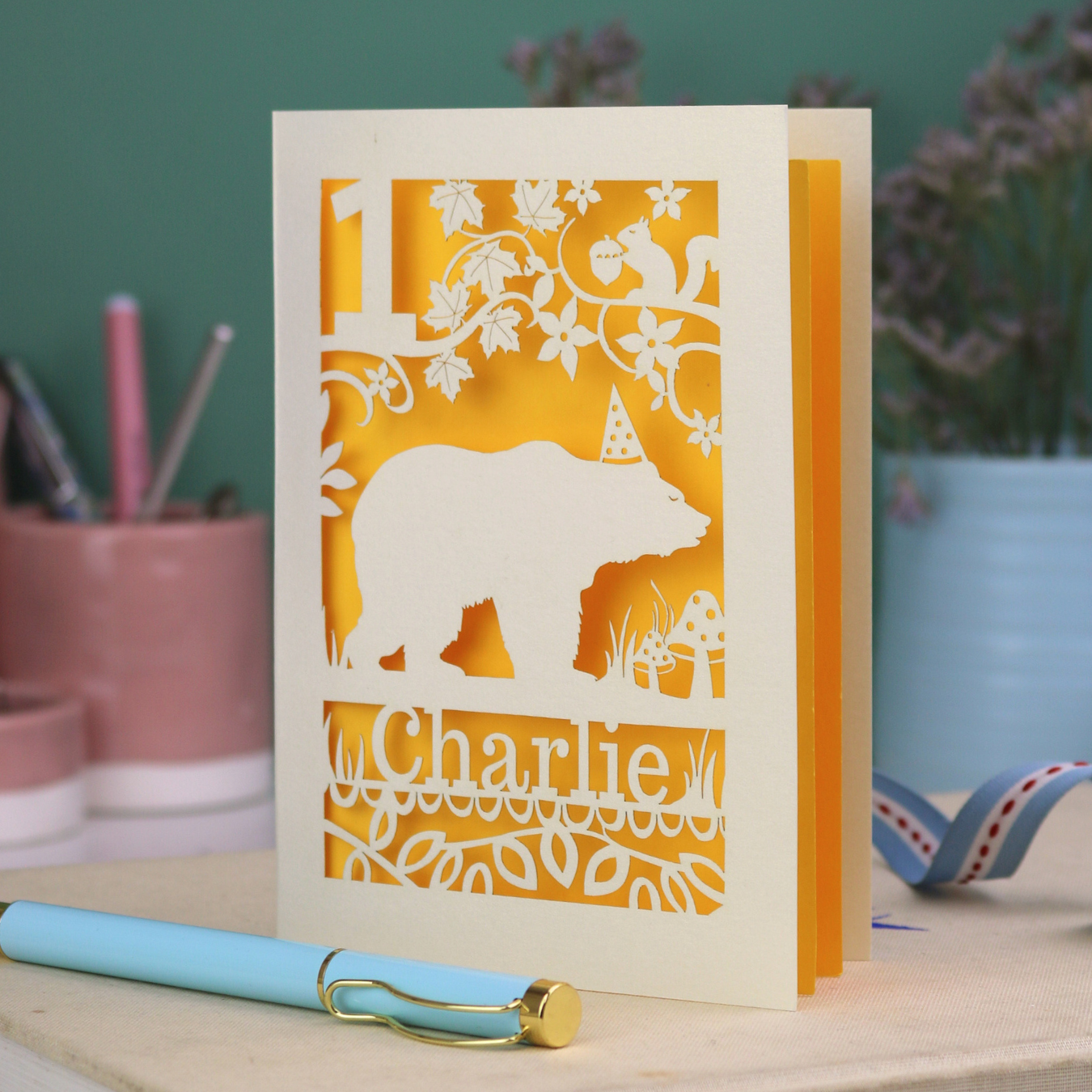 A laser cut birthday card personalised with an age and name, and featuring a cut out bear in a party hat with woodland border.