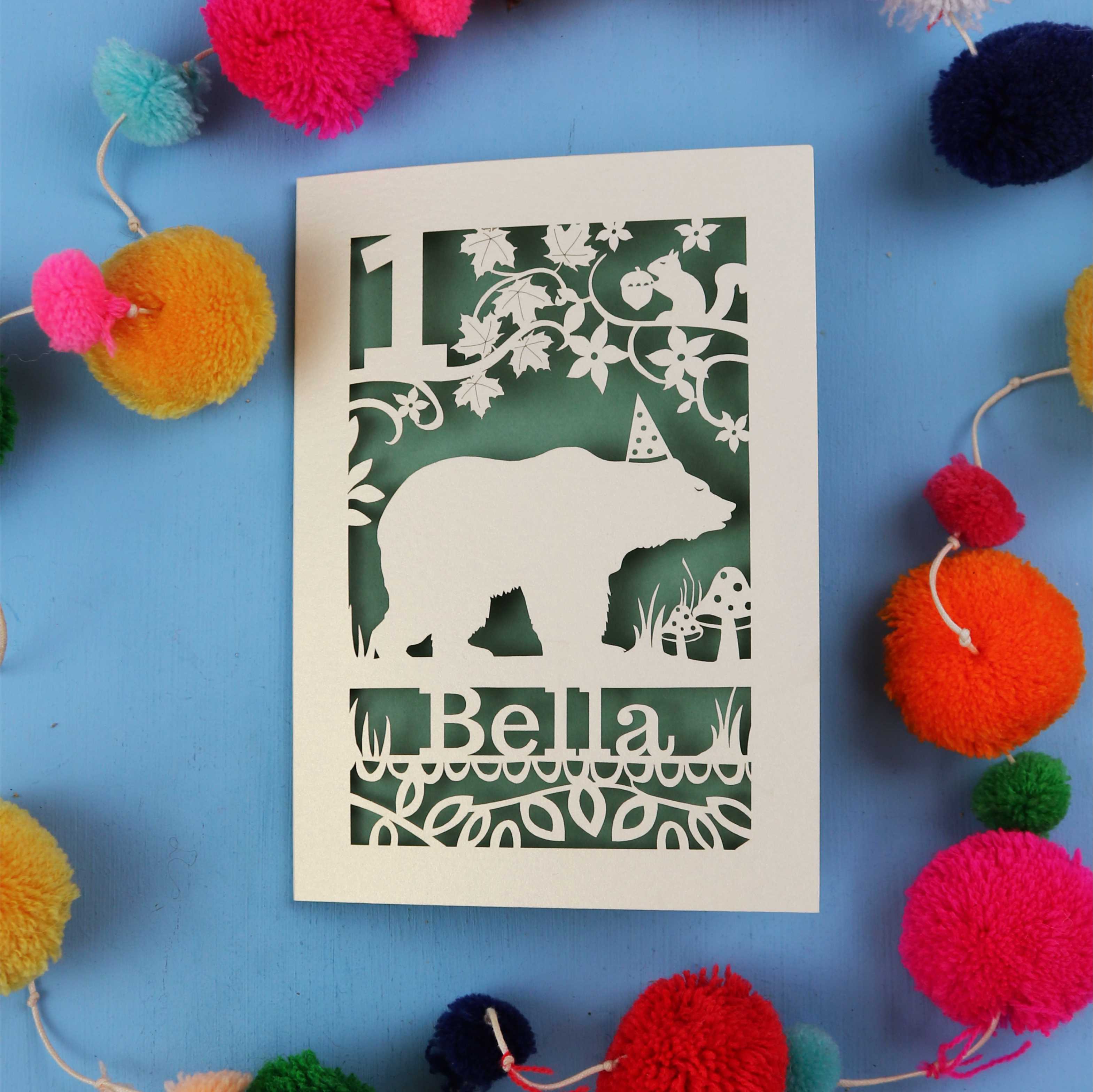 1st birthday Card with bear design and 'Bella' text on a blue background with colourful pom-poms.