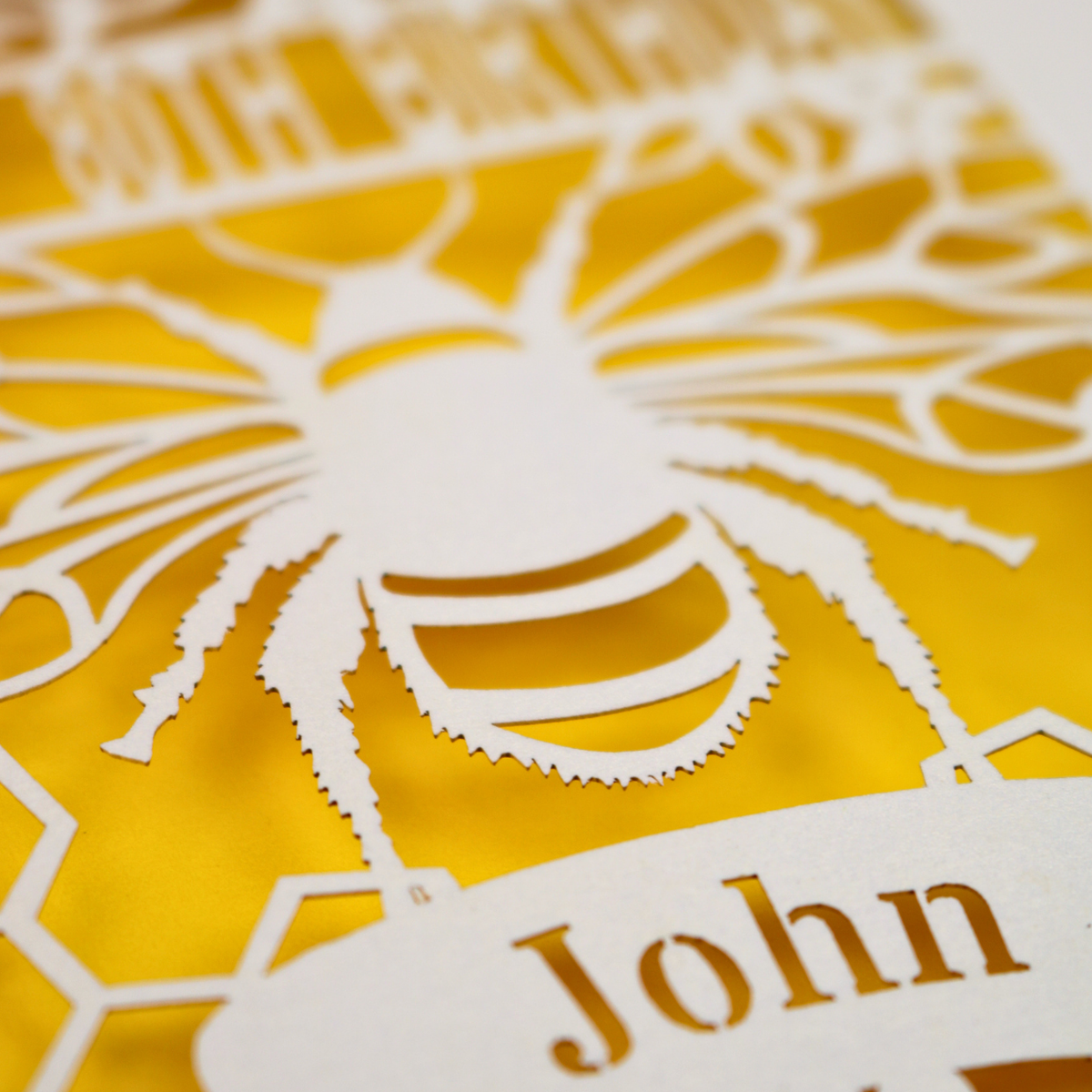 Close-up of a laser-cut personalized birthday card featuring a bee design over a bright yellow background, with the name 'John' at the bottom.