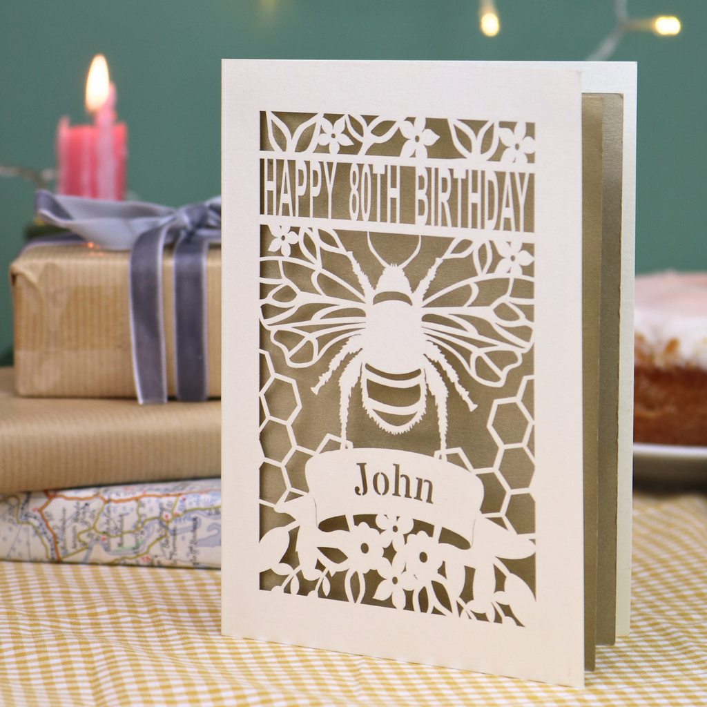 A personalized laser-cut 80th birthday card with a bee and honeycomb design, featuring the text 'HAPPY 80TH BIRTHDAY' and 'John', displayed on a checked tablecloth with a blurred birthday cake and wrapped gifts in the background.