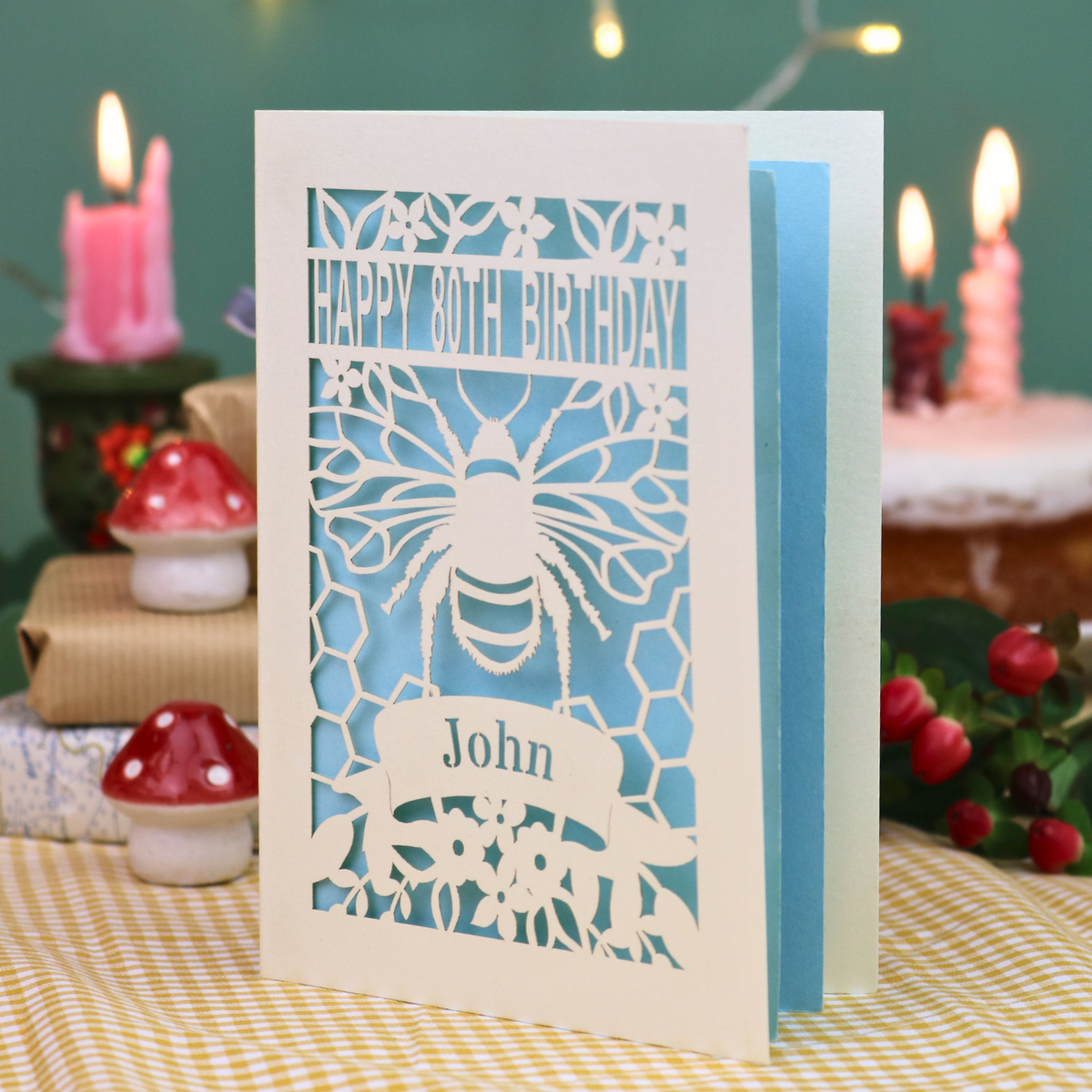 A personalised 80th birthday card with a bee and honeycomb design, featuring the text 'HAPPY 80TH BIRTHDAY' and 'John', displayed on a checked tablecloth with a blurred birthday cake and wrapped gifts in the background.