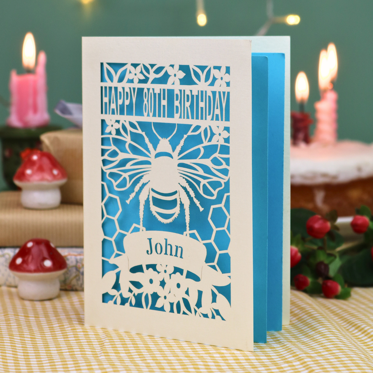 A personalised 80th birthday card with a bee and honeycomb design, featuring the text 'HAPPY 80TH BIRTHDAY' and 'John', displayed on a checked tablecloth with a blurred birthday cake and wrapped gifts in the background.