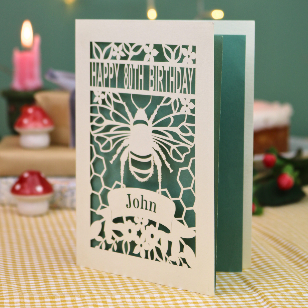 A personalised 80th birthday card with a bee and honeycomb design, featuring the text 'HAPPY 80TH BIRTHDAY' and 'John', displayed on a checked tablecloth with a blurred birthday cake and wrapped gifts in the background.