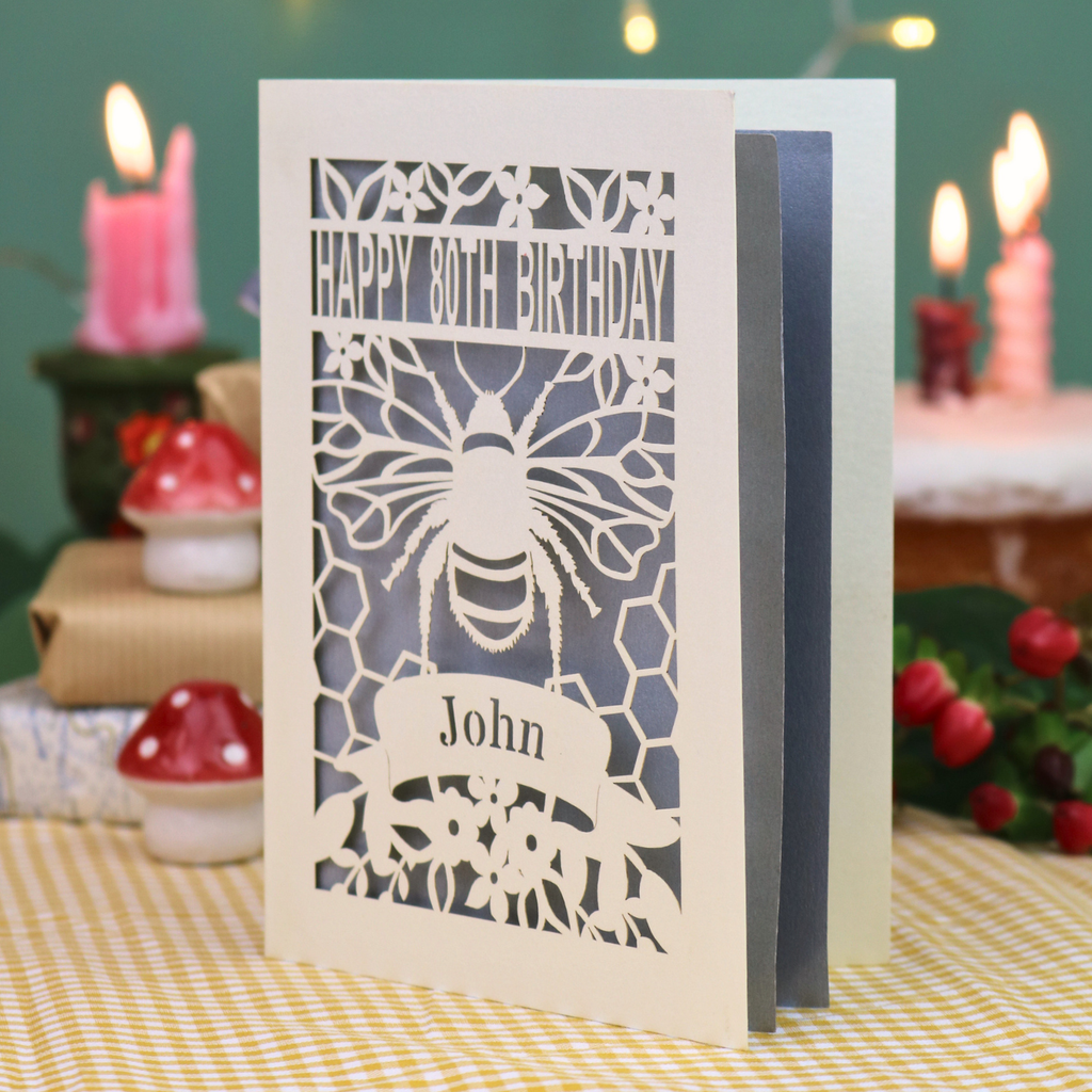 A personalised 80th birthday card with a bee and honeycomb design, featuring the text 'HAPPY 80TH BIRTHDAY' and 'John', displayed on a checked tablecloth with a blurred birthday cake and wrapped gifts in the background.