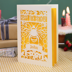 A personalized laser-cut 80th birthday card with a bee and honeycomb design, featuring the text 'HAPPY 80TH BIRTHDAY' and 'John', displayed on a checked tablecloth with a blurred birthday cake and wrapped gifts in the background.