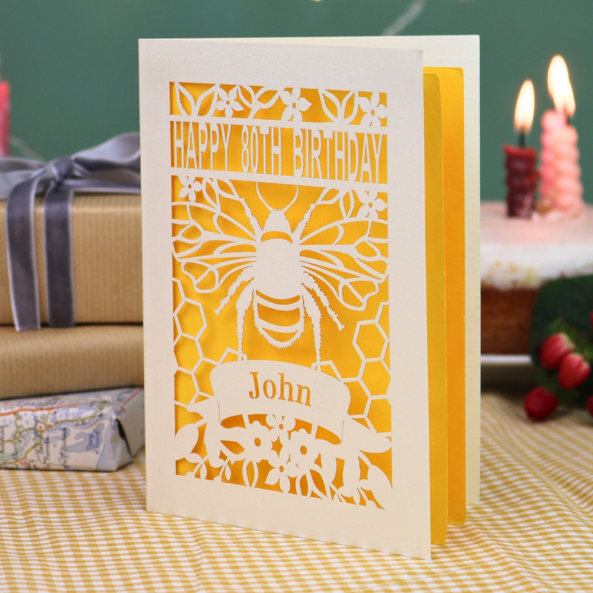 A personalized laser-cut 80th birthday card with a bee and honeycomb design, featuring the text 'HAPPY 80TH BIRTHDAY' and 'John', displayed on a checked tablecloth with a blurred birthday cake and wrapped gifts in the background.