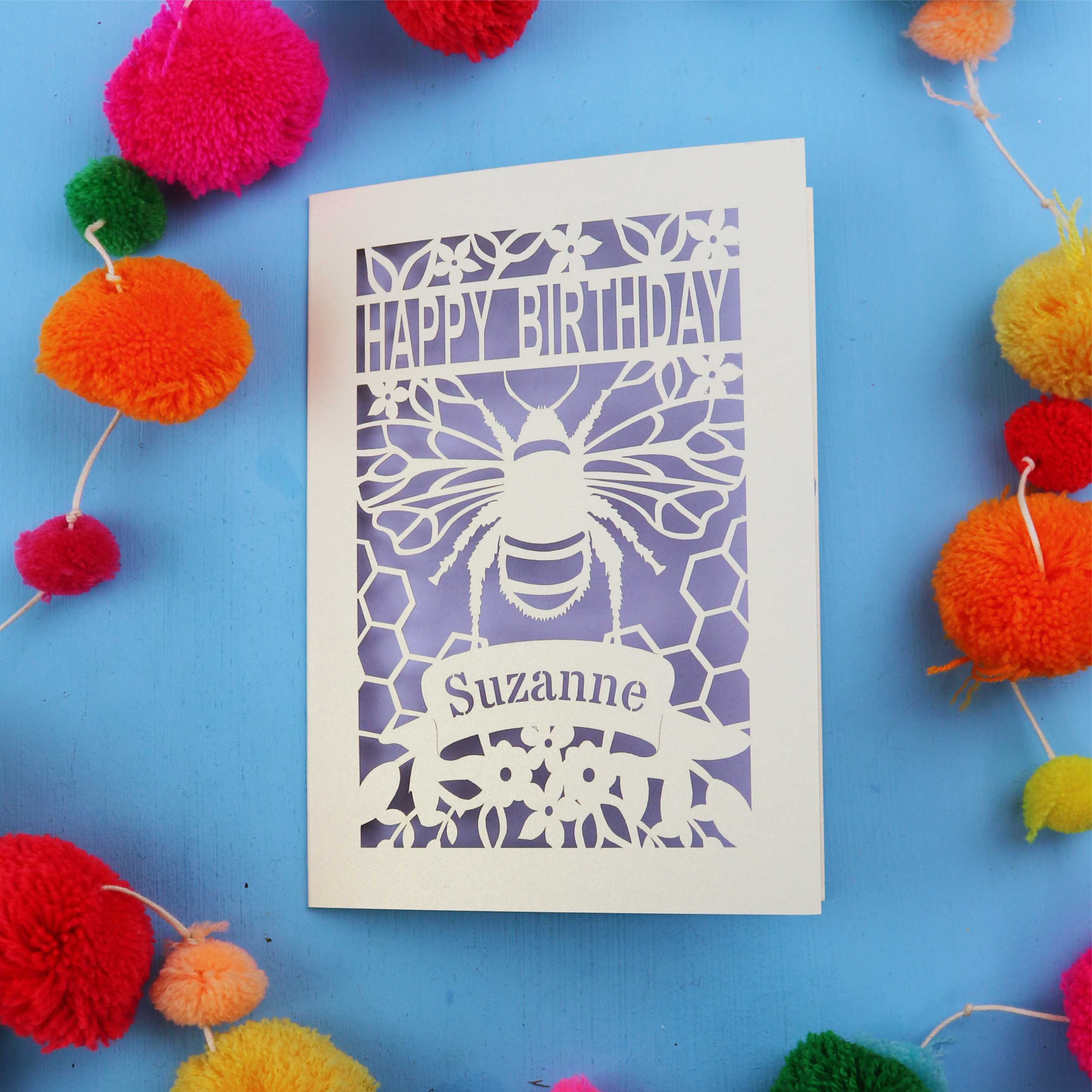 Laser cut Birthday card with bee design and colourful pom-poms on a blue background