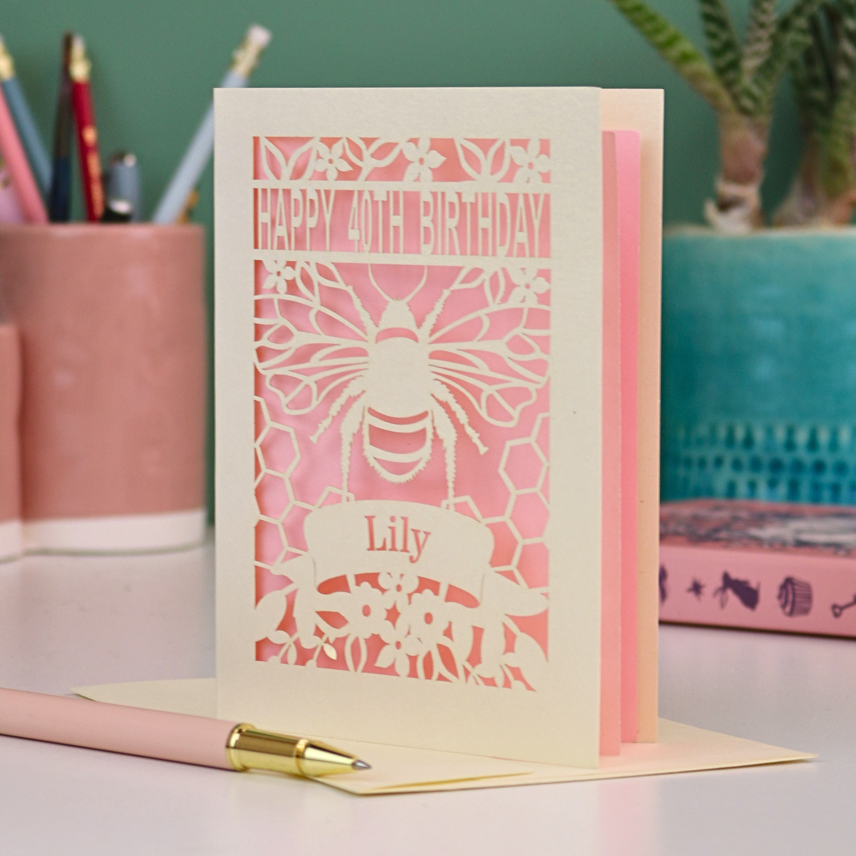 Laser-cut birthday card with cut out bee and honeycomb design and 'Happy 40th Birthday' text, personalised with the recipient's name in a banner underneath, on a desk.