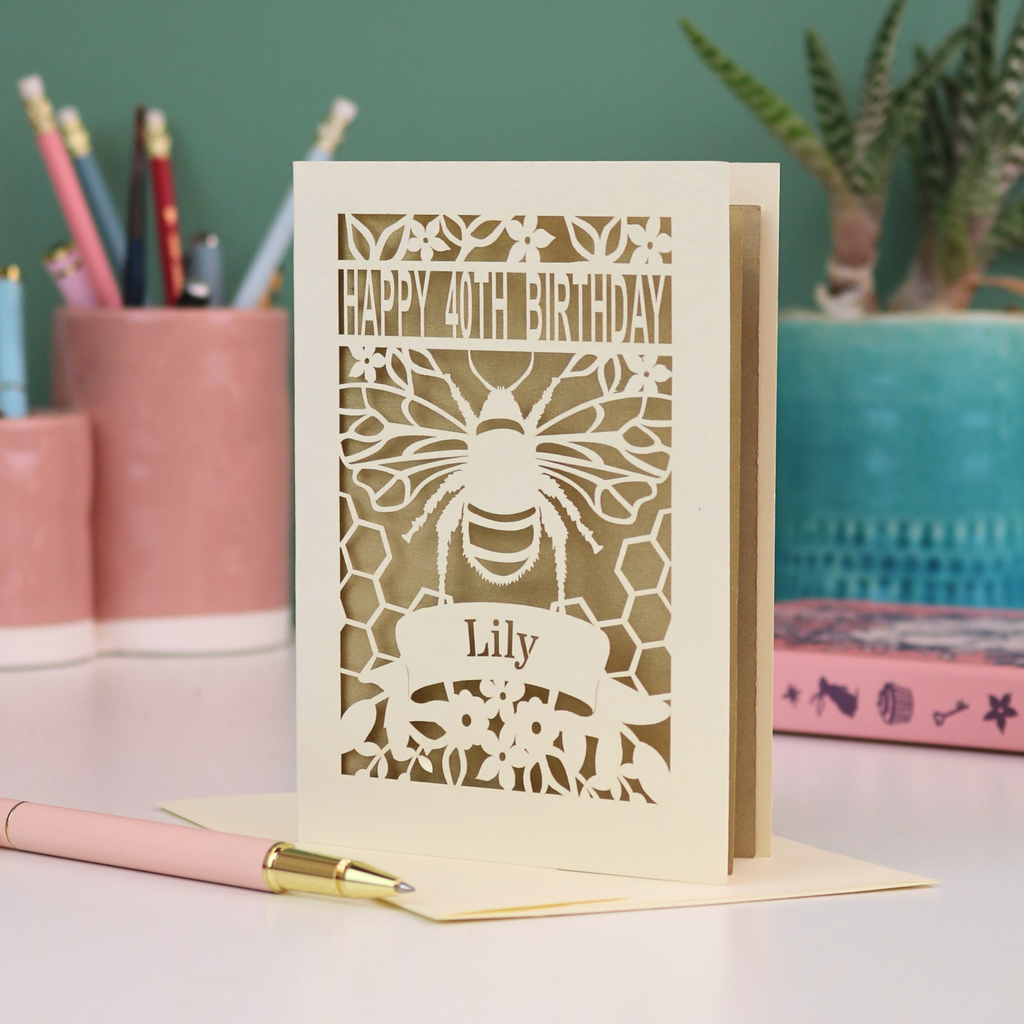Laser-cut birthday card with cut out bee and honeycomb design and 'Happy 40th Birthday' text, personalised with the recipient's name in a banner underneath, on a desk.