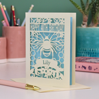 Laser-cut birthday card with cut out bee and honeycomb design and 'Happy 40th Birthday' text, personalised with the recipient's name in a banner underneath, on a desk.