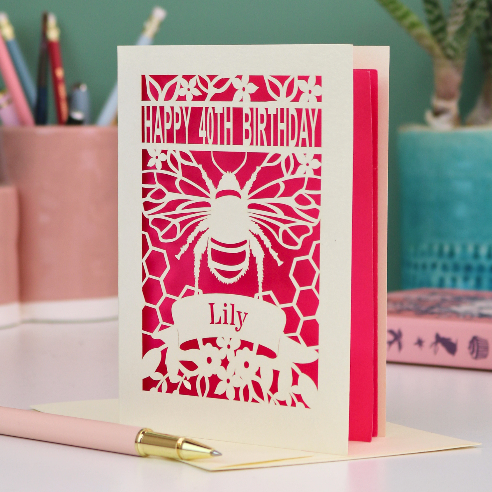 Laser-cut birthday card with cut out bee and honeycomb design and 'Happy 40th Birthday' text, personalised with the recipient's name in a banner underneath, on a desk.