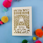 Laser-cut personalised Valentines card with 'Bee Mine' and 'Helen' text on a blue background with colourful pom-poms.