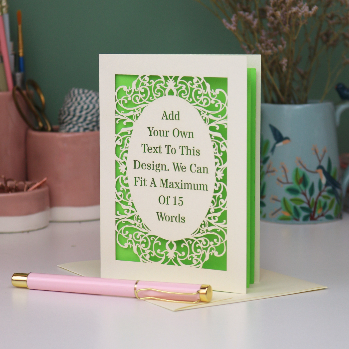 A custom laser cut personalised greeting card with a cream ornate filigree border and a green backing, displayed on a white surface with a pink pen in the foreground. The card features placeholder text that reads 'Add Your Own Text To This Design.