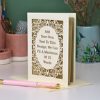 A custom laser-cut greeting card with a cream and gold design, displayed on a white surface with a pink pen in the foreground and decorative background items.