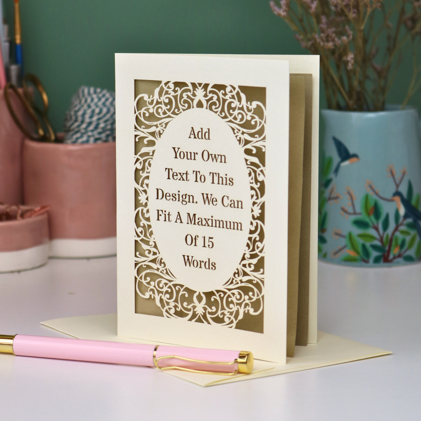 A custom laser-cut greeting card with a cream and gold design, displayed on a white surface with a pink pen in the foreground and decorative background items.
