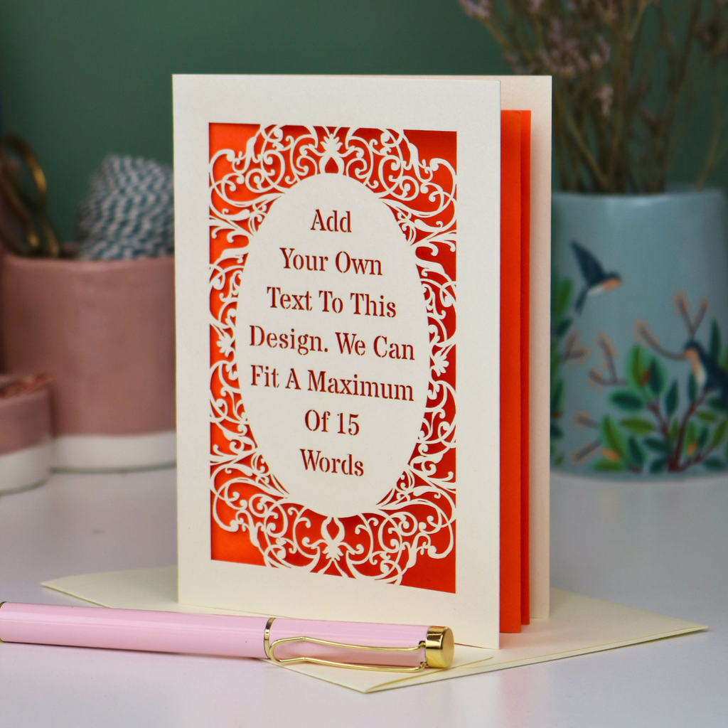 A bespoke cut out personalised greeting card with a cream ornate filigree border and orange backing, displayed on a white surface with a pink pen in the foreground. The card features placeholder text that reads 'Add Your Own Text To This Design.