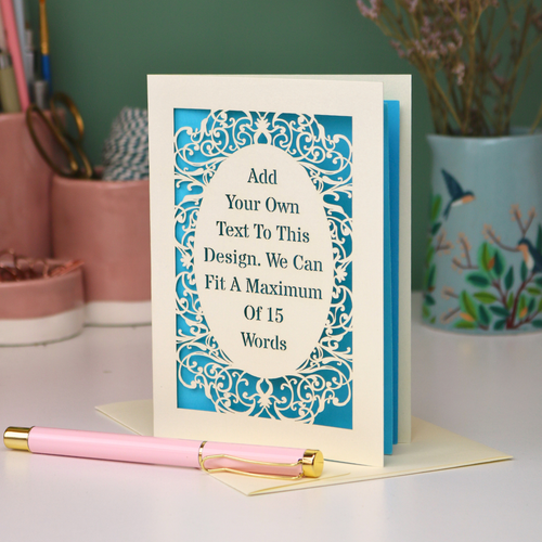 A bespoke personalised greeting card with a cream ornate filigree border and a turquoise blue backing, displayed on a white surface with a pink pen in the foreground. The card features placeholder text that reads 'Add Your Own Text To This Design.