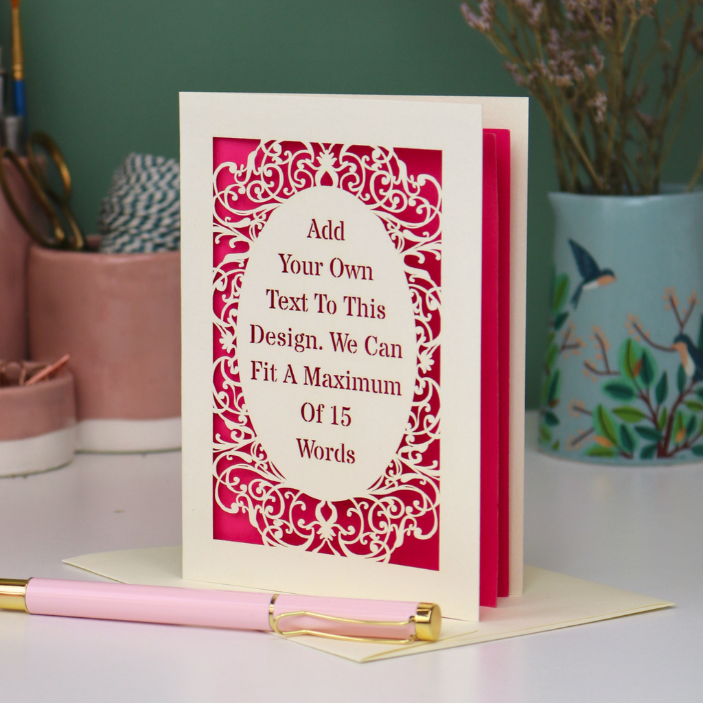 A bespoke personalised greeting card with a cream ornate filigree border and a shocking pink backing, displayed on a white surface with a pink pen in the foreground. The card features placeholder text that reads 'Add Your Own Text To This Design.