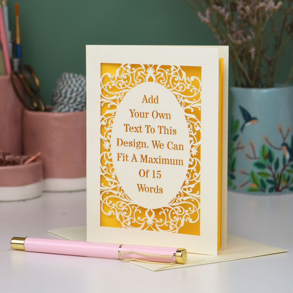 A bespoke laser cut personalised greeting card with a cream ornate filigree border and a yellow backing, displayed on a white surface with a pink pen in the foreground. The card features placeholder text that reads 'Add Your Own Text To This Design.