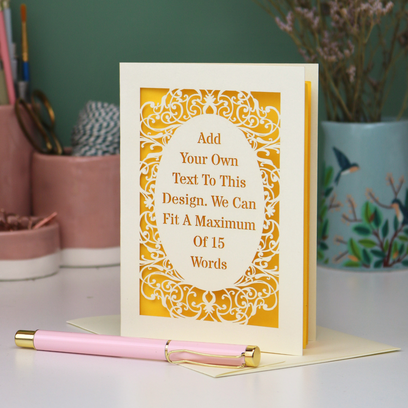 A bespoke laser cut personalised greeting card with a cream ornate filigree border and a yellow backing, displayed on a white surface with a pink pen in the foreground. The card features placeholder text that reads 'Add Your Own Text To This Design.