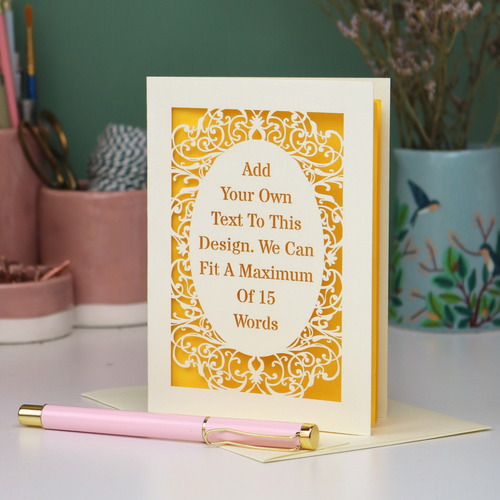 A bespoke laser cut personalised greeting card with a cream ornate filigree border and a yellow backing, displayed on a white surface with a pink pen in the foreground. The card features placeholder text that reads 'Add Your Own Text To This Design.