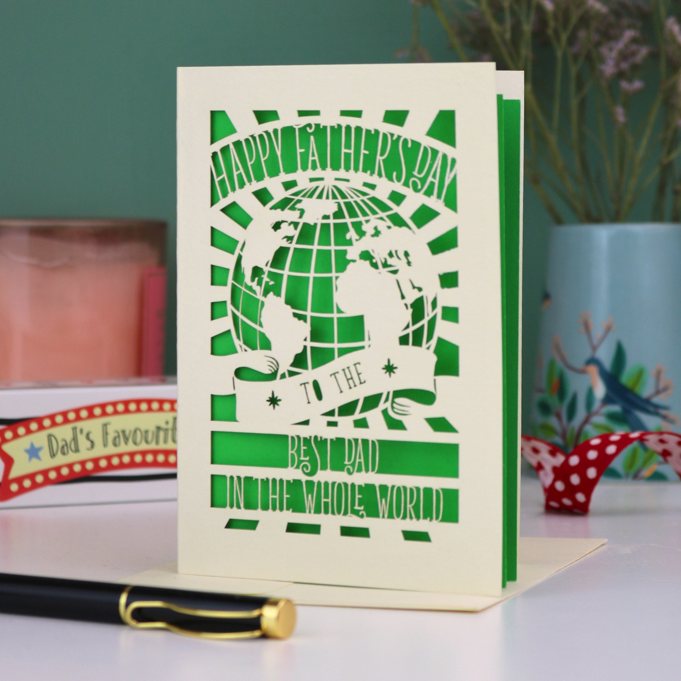 A laser cut Father's Day card with globe design and the text "Happy Father's Day to the Best Dad In the Whole world", on a desk with pen and stationery items