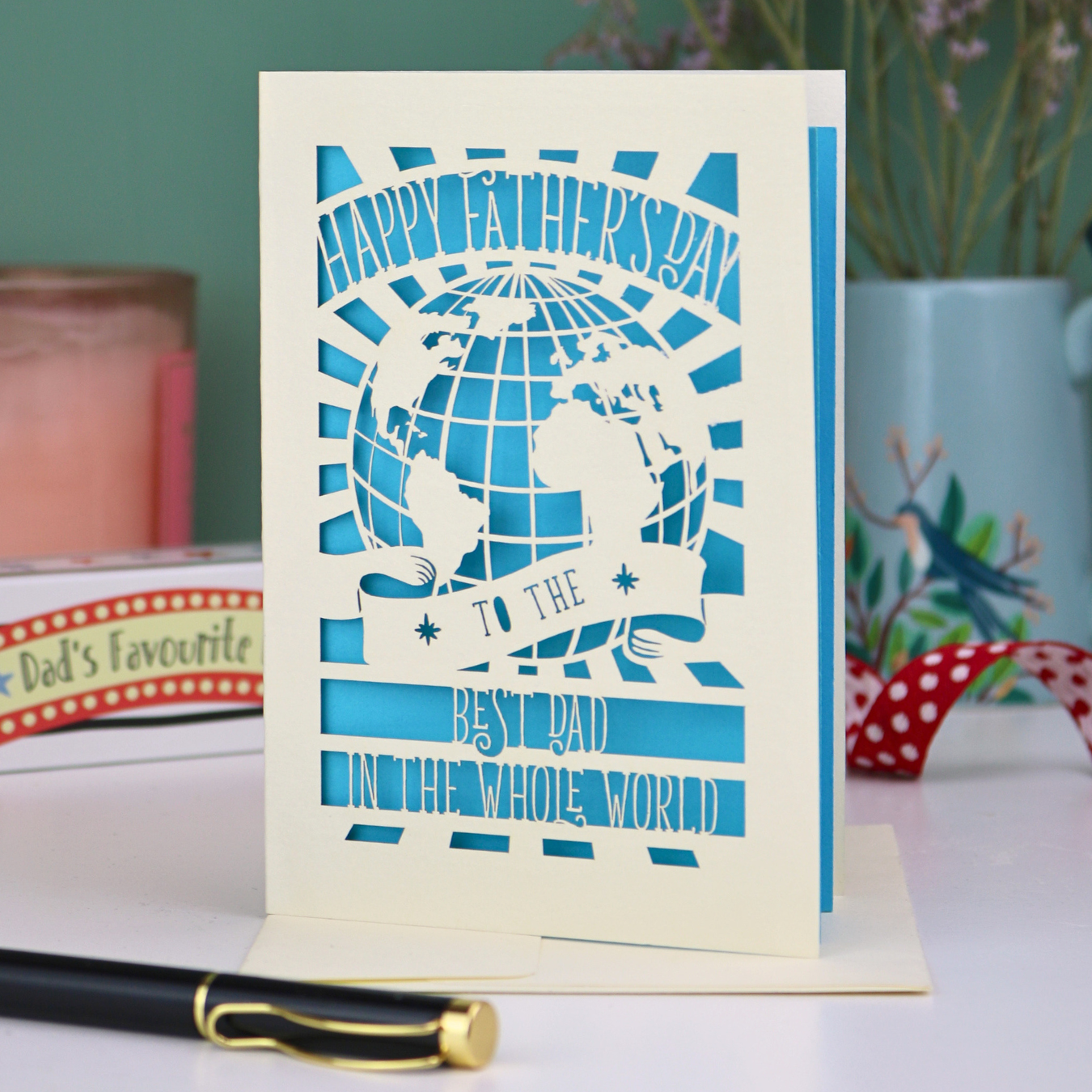 A unique laser cut Father's Day card with globe design and the text "Happy Father's Day to the Best Dad In the Whole world", on a desk with pen and stationery items