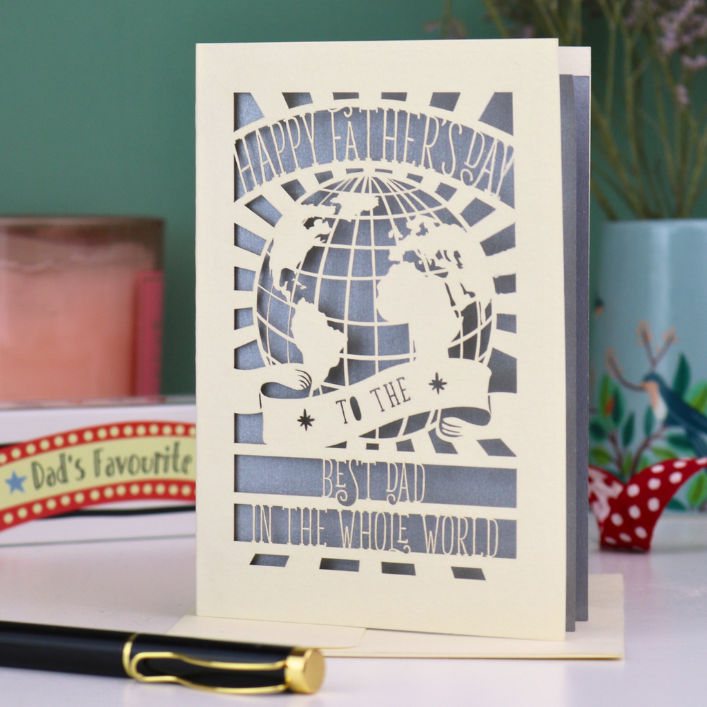 A paper cut Father's Day card with globe design and the text "Happy Father's Day to the Best Dad In the Whole world", on a desk with pen and stationery items