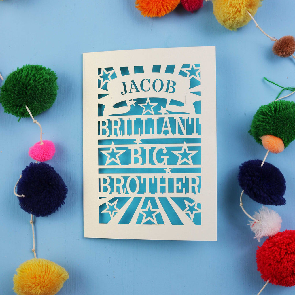 Laser cut greetings card with 'Jacob Brilliant Big Brother' text on a blue background with colourful pom-poms.