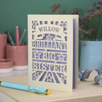 Card with 'Willow, Brilliant Big Sister' text on a desk with stationery items.