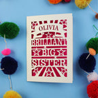 Card with 'Olivia Brilliant Big Sister' text on a blue background with colouful pom-poms.