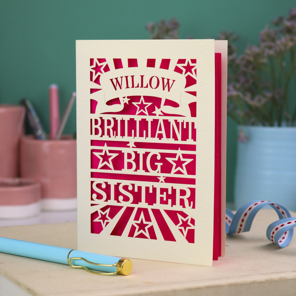 Card with 'WILLOW BRILLIANT BIG SISTER' text and star patterns on a desk with stationery items.