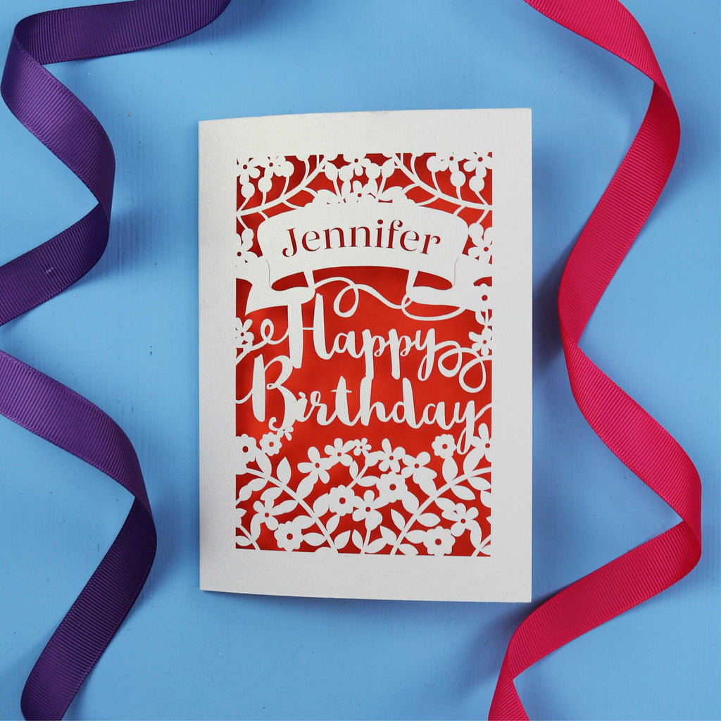A laser cut birthday card with the recipient's name laser cut from a banner shape and the words Happy Birthday in the middle, surrounded by floral designs. Birthday card is cream with orange paper behind