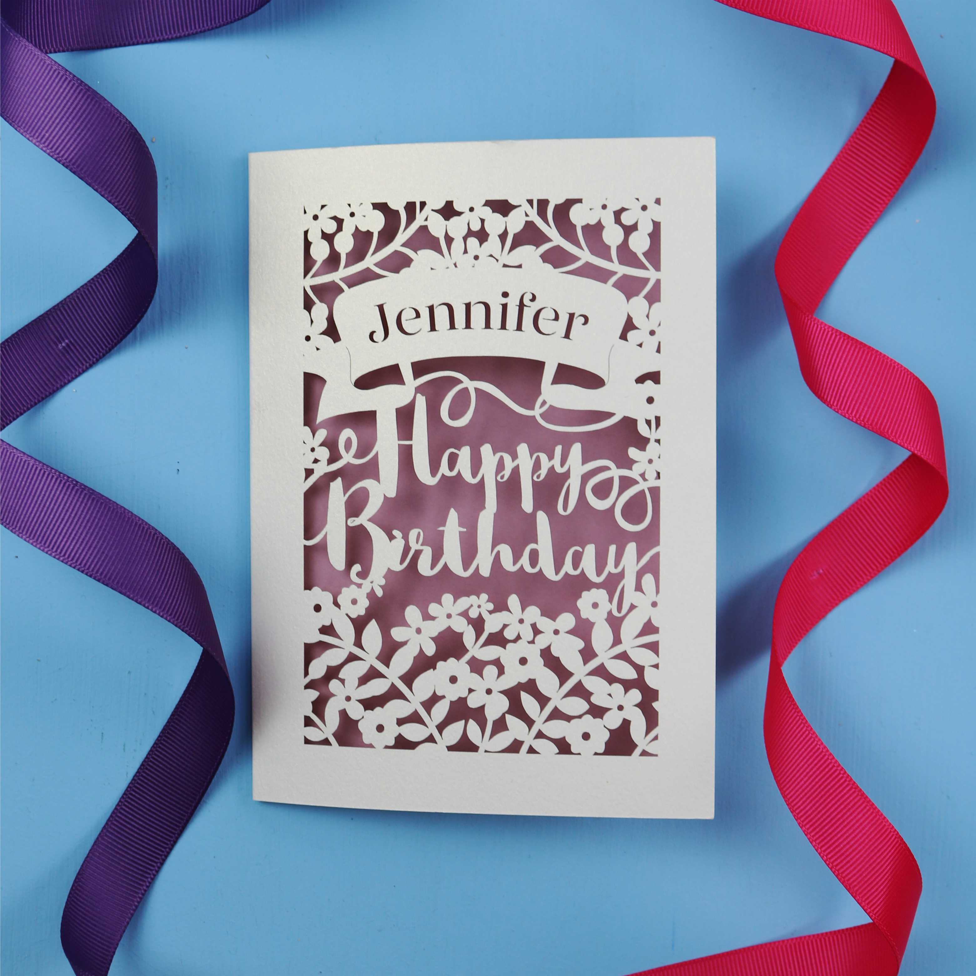 A cut out birthday card with the recipient's name laser cut from a banner shape and the words Happy Birthday in the middle, surrounded by floral designs. Birthday card is cream with dusky pink paper behind
