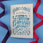 A unique birthday card with the recipient's name laser cut from a banner shape and the words Happy Birthday in the middle, surrounded by floral designs. Birthday card is cream with light blue paper behind