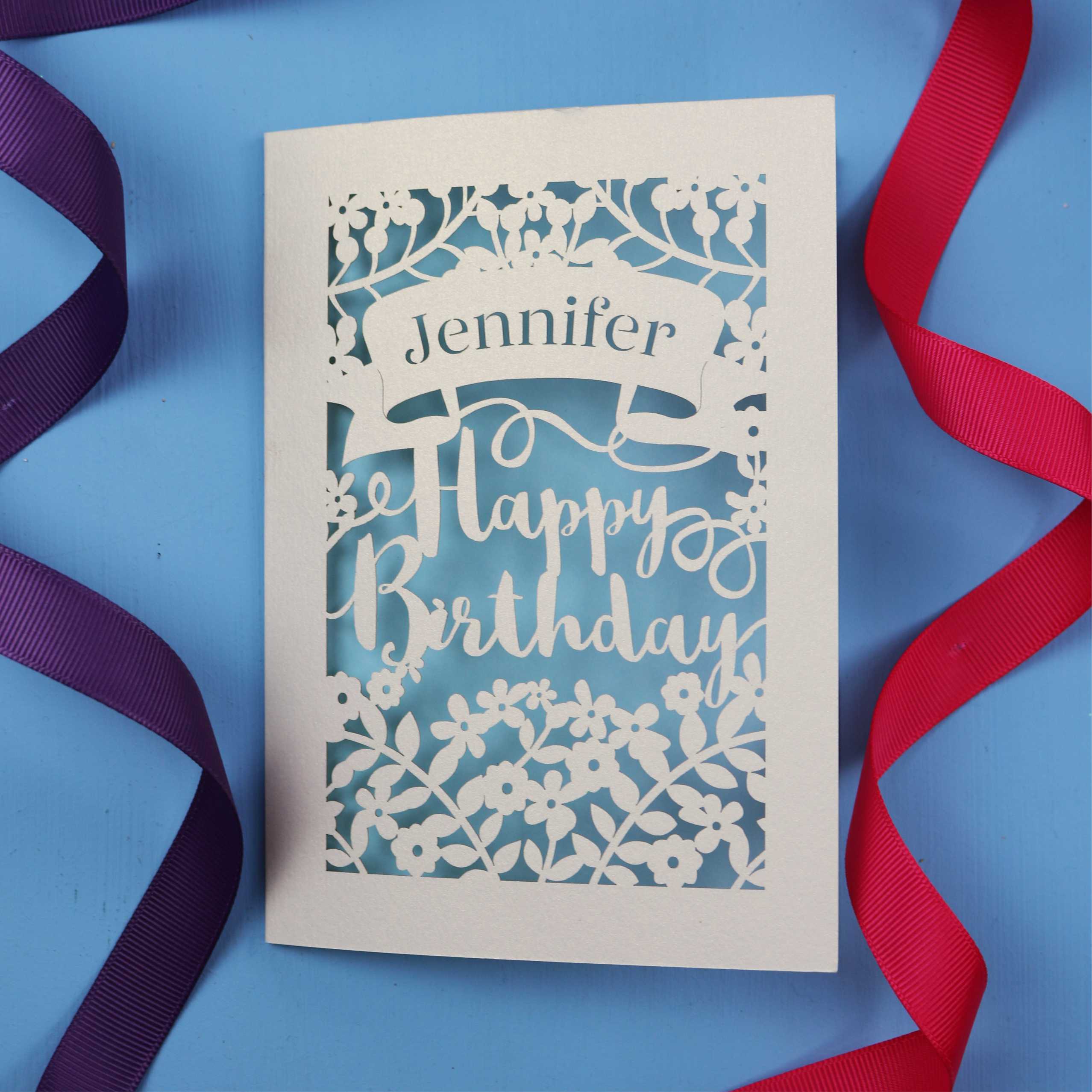 A unique birthday card with the recipient's name laser cut from a banner shape and the words Happy Birthday in the middle, surrounded by floral designs. Birthday card is cream with light blue paper behind