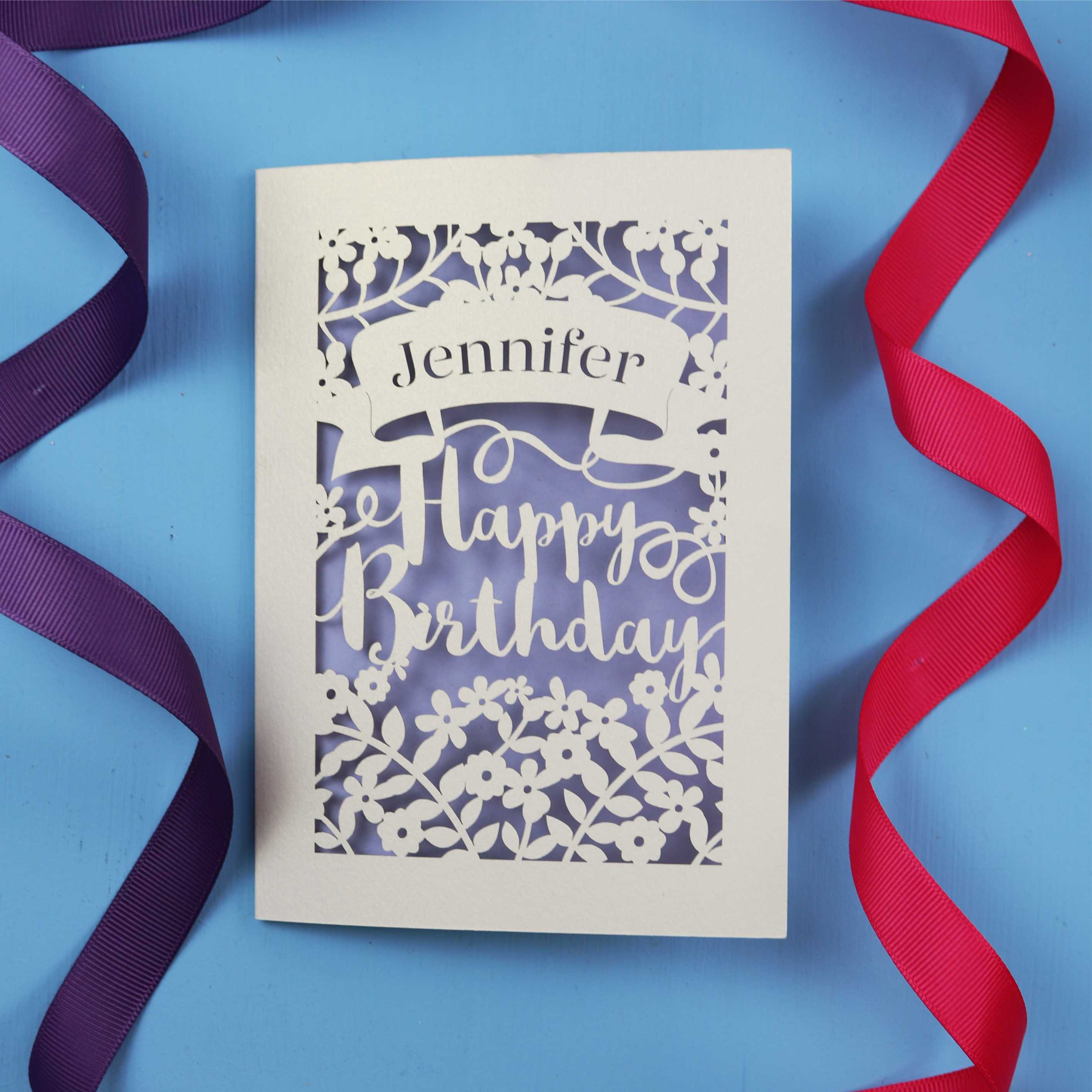 A special birthday card with the recipient's name laser cut from a banner shape and the words Happy Birthday in the middle, surrounded by floral designs. Birthday card is cream with lilac paper behind