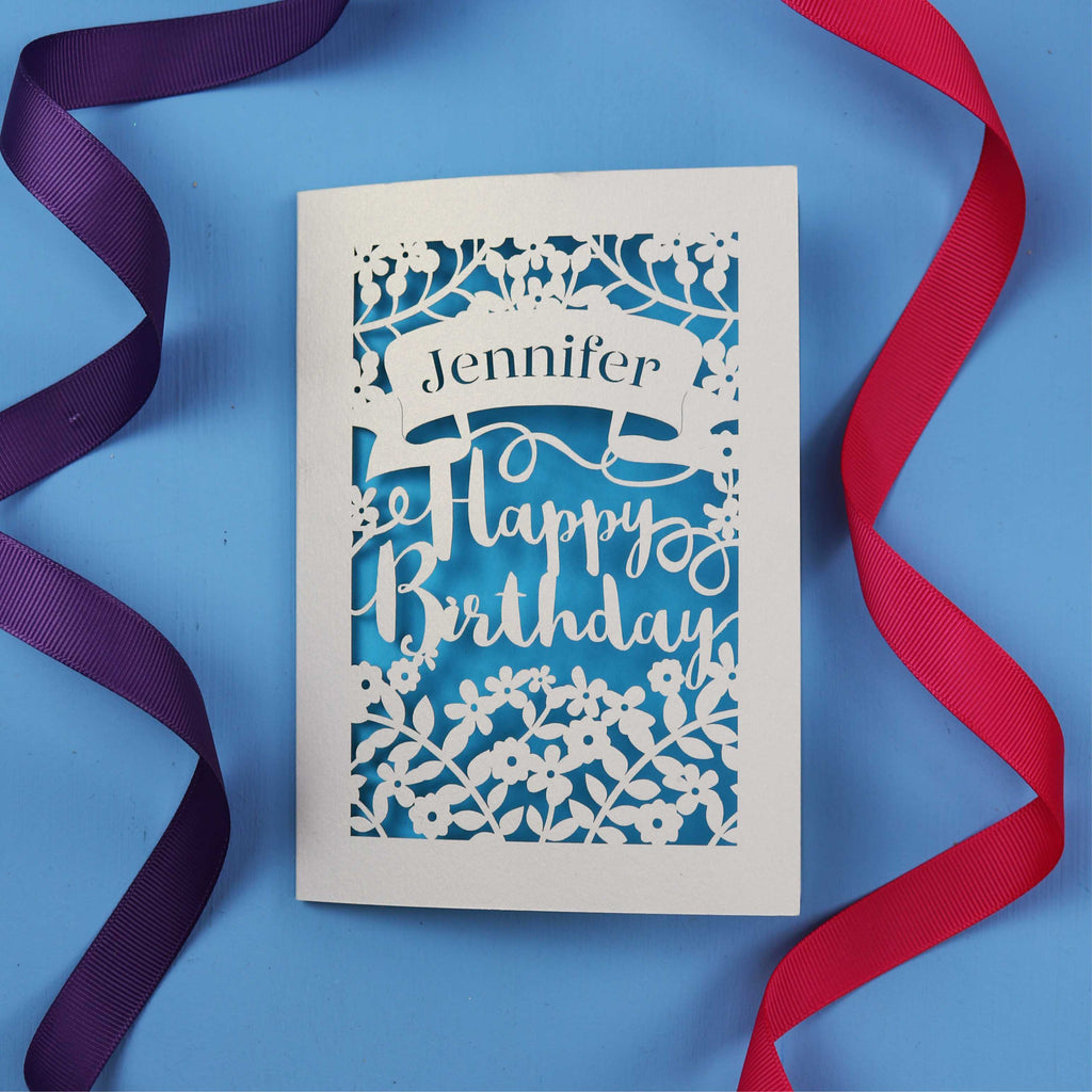 A UK made birthday card with the recipient's name laser cut from a banner shape and the words Happy Birthday in the middle, surrounded by floral designs. Birthday card is cream with blue paper behind