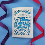 A UK made birthday card with the recipient's name laser cut from a banner shape and the words Happy Birthday in the middle, surrounded by floral designs. Birthday card is cream with blue paper behind