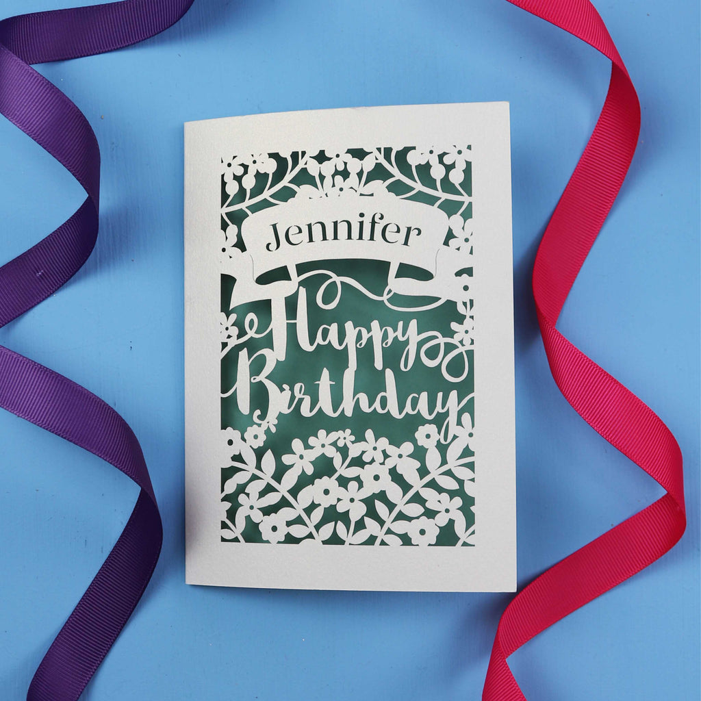 A personalised birthday card with the recipient's name laser cut from a banner shape and the words Happy Birthday in the middle, surrounded by floral designs. Birthday card is cream with sage paper behind