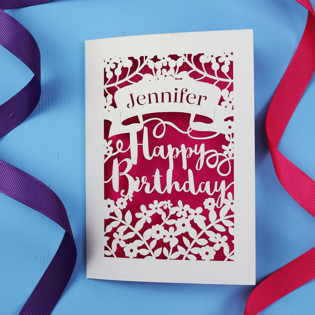 A personalised birthday card with the recipient's name laser cut from a banner shape and the words Happy Birthday in the middle, surrounded by floral designs. Birthday card is cream with shocking pink paper behind