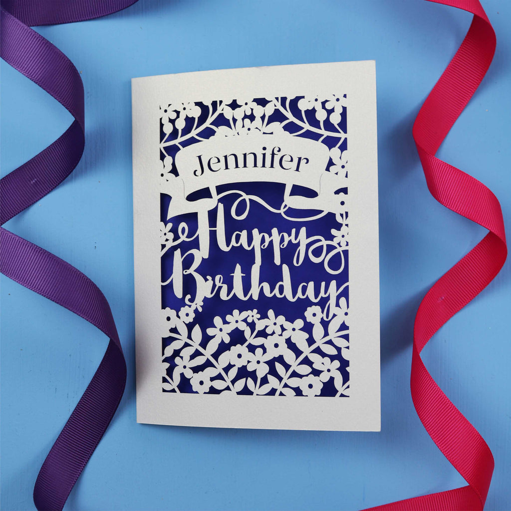A personalised birthday card with the recipient's name laser cut from a banner shape and the words Happy Birthday in the middle, surrounded by floral designs. Birthday card is cream with violet paper behind