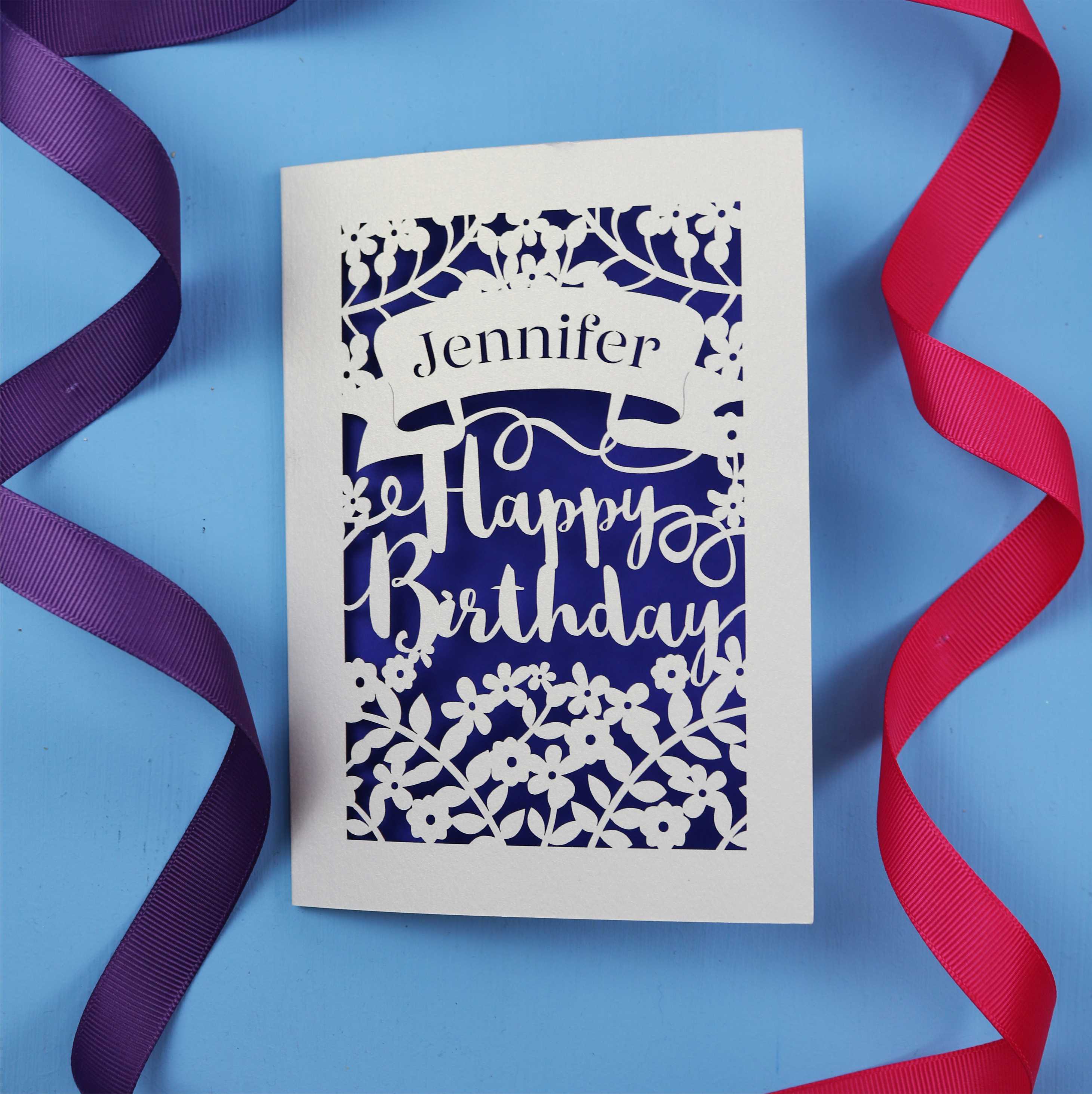 A personalised birthday card with the recipient's name laser cut from a banner shape and the words Happy Birthday in the middle, surrounded by floral designs. Birthday card is cream with violet paper behind