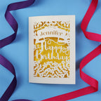 A personalised birthday card with the recipient's name laser cut from a banner shape and the words Happy Birthday in the middle, surrounded by floral designs. Birthday card is cream with yellow paper behind