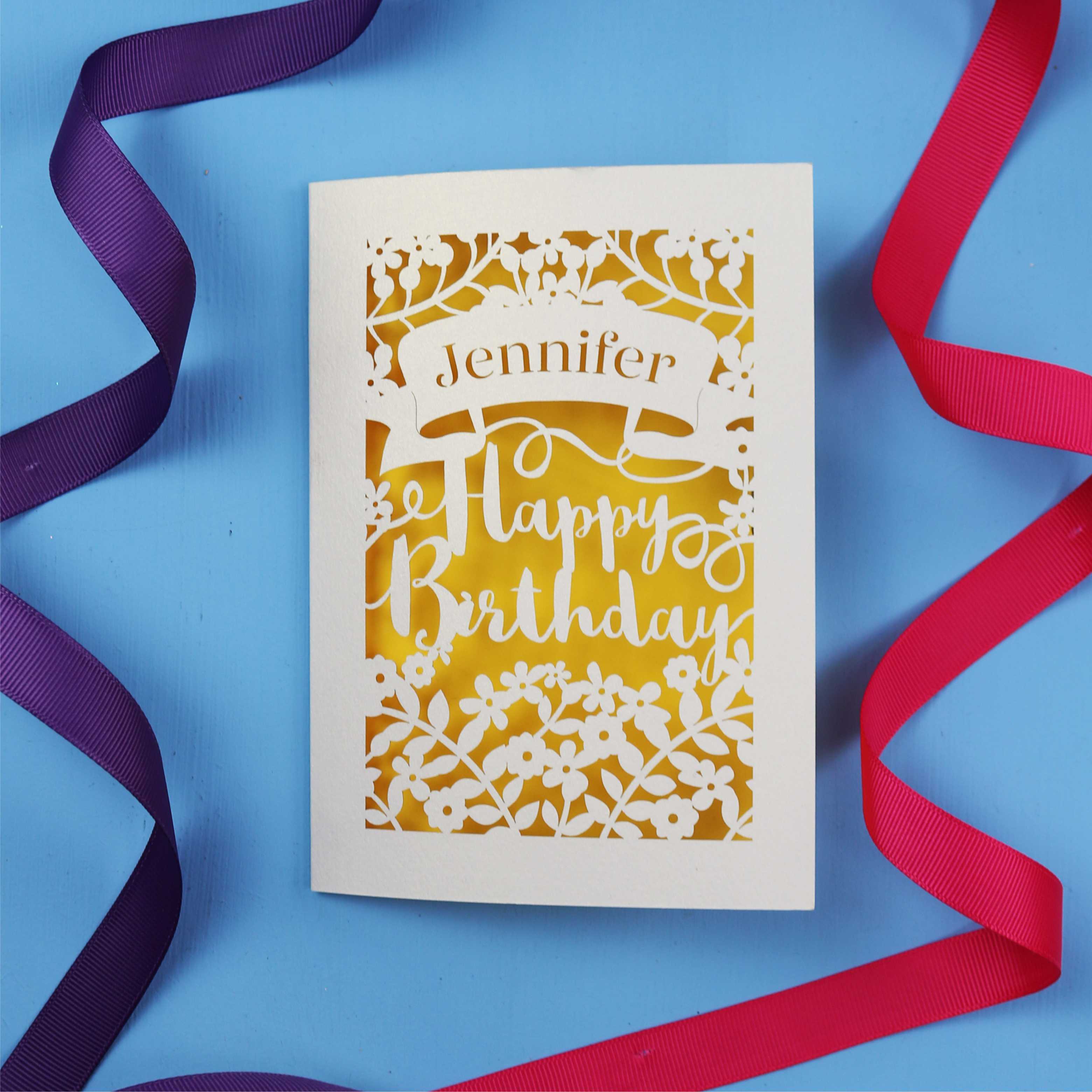 A personalised birthday card with the recipient's name laser cut from a banner shape and the words Happy Birthday in the middle, surrounded by floral designs. Birthday card is cream with yellow paper behind